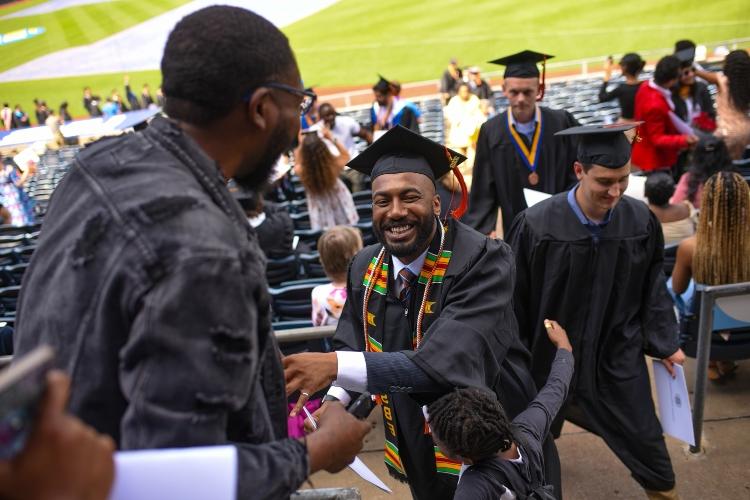 A graduate walks up the stairs after receiving his diploma and shakes hands with a loved one as a young boy hugs him